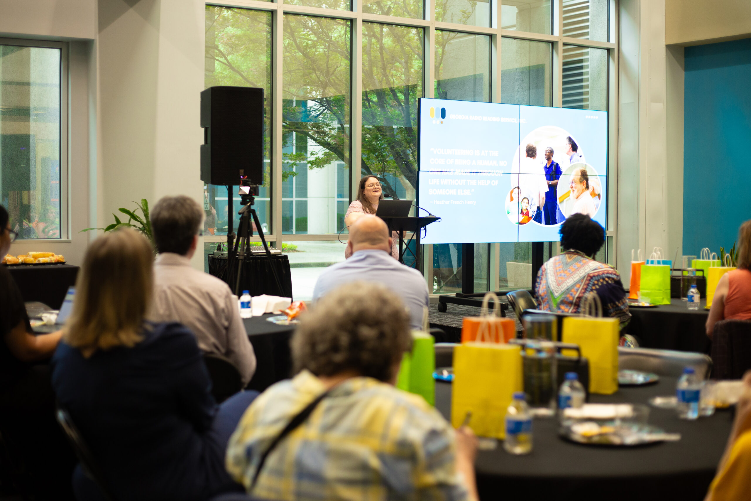 A Caucasian Woman Wearing A Pink Floral Dress Stands At A Podium Speaking During A Volunteer Appreciation Event Held By Georgia Radio Reading Service, Inc. She Is Addressing An Attentive Audience Seated At Round Tables Covered With Black Tablecloths And Decorated With Colorful Gift Bags, Snacks, And Water Bottles. Behind The Speaker Is A Large Screen Displaying A Quote About Volunteering And Images Of People Engaged In Community Service. Large Windows Provide Natural Light, Revealing Trees And A Modern Building Exterior. The Atmosphere Is Warm And Engaging, Celebrating The Contributions Of Volunteers.