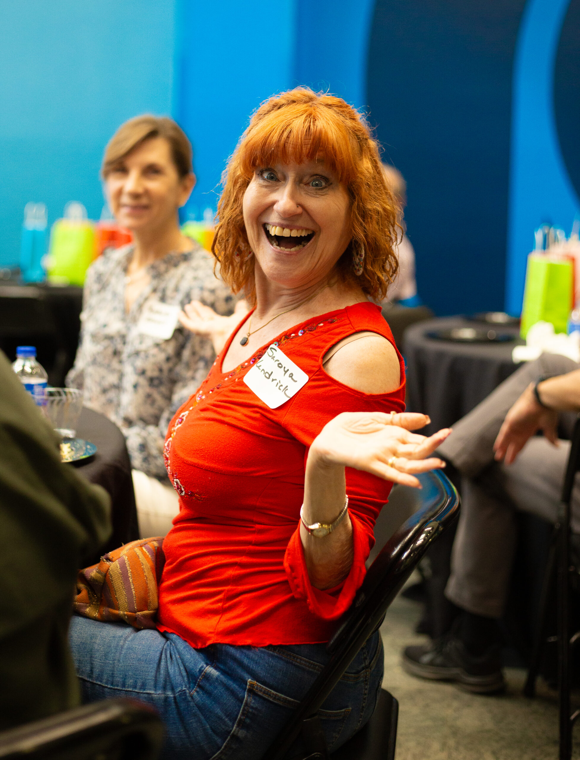This Image Shows A Joyful Woman With Red Curly Hair Sitting At A Round Table During What Appears To Be A Social Or Professional Event. She Is Wearing A Vibrant Red Top With Cut-Out Shoulders And Blue Jeans. Her Name Tag Reads &Quot;Sonya&Quot;, And She Is Smiling Brightly While Holding Up A Small Gift Card Or Voucher That Says &Quot;Be Our Guest&Quot; From Chick-Fil-A. Her Excitement And Enthusiasm Are Clearly Visible In Her Expression.  In The Background, Other People Are Seated At Tables, Suggesting A Gathering Or Celebration. The Setting Includes Colorful Gift Bags, Drinks, And Decorations, Contributing To A Festive And Welcoming Atmosphere. The Walls Are Painted In Shades Of Blue, Adding To The Cheerful Ambiance.