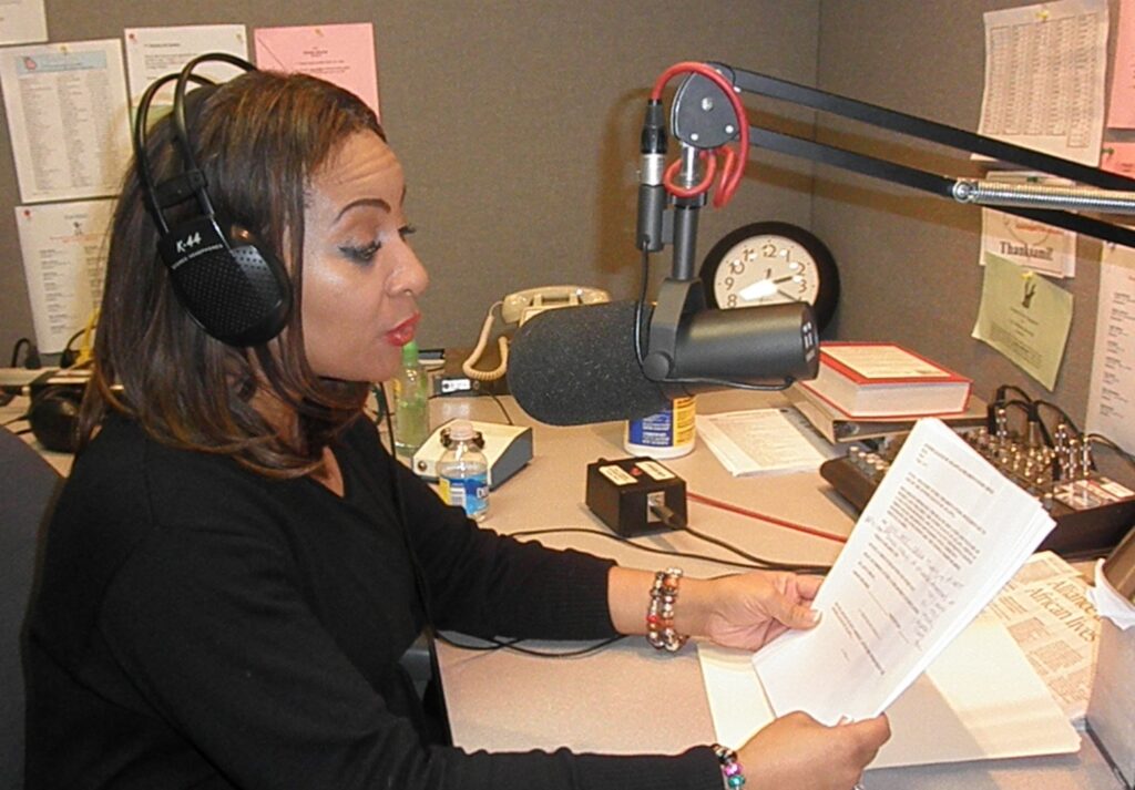 A Woman Sits In A Radio Studio Wearing Large Headphones And Speaking Into A Professional Microphone. She Holds Printed Papers While Reading Aloud. Behind Her Are A Wall Clock, Sound Equipment, And Various Papers Pinned To The Cubicle Wall.