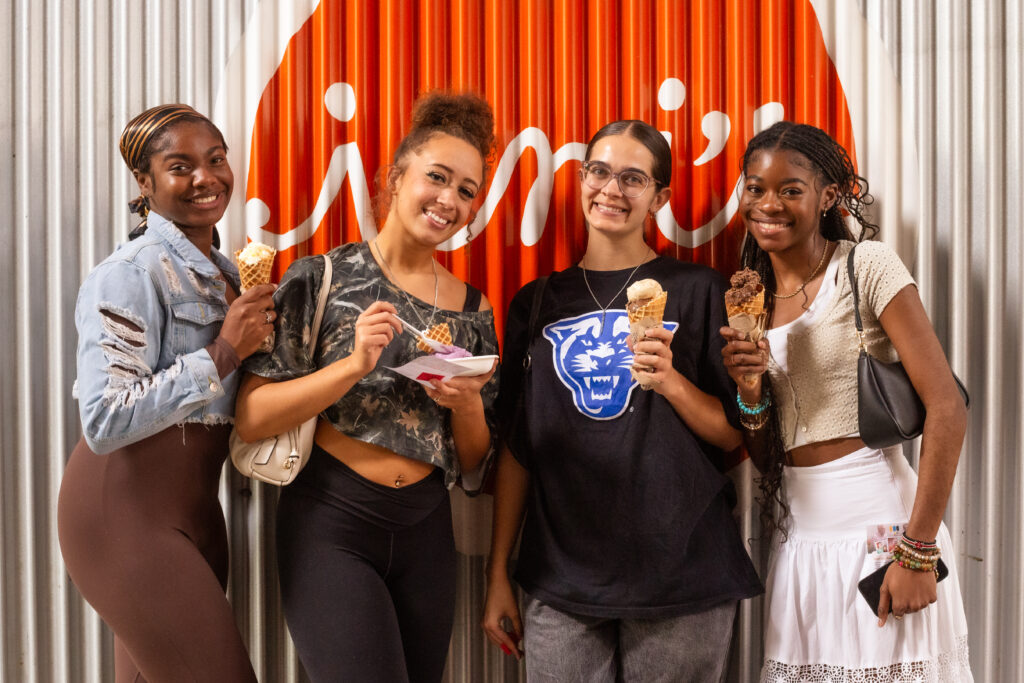 Four Young Women Stand Together In Front Of A Corrugated Metal Wall With An Orange And White Logo. They Are Smiling And Holding Ice Cream Cones, With One Woman Holding A Bowl. Each Is Casually Dressed In Different Styles, Including A Denim Jacket, Crop Top, Graphic T-Shirt, And White Skirt.