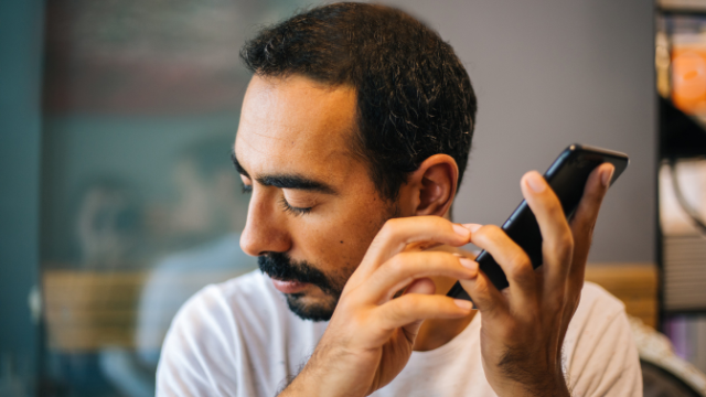 A Man With Short Dark Hair And A Beard Holds A Smartphone In His Hand While Touching The Screen With His Other Hand. His Eyes Are Closed, And He Appears To Be Using The Device By Feel Or Voice Command. The Background Is Softly Blurred, Keeping Focus On The Man.