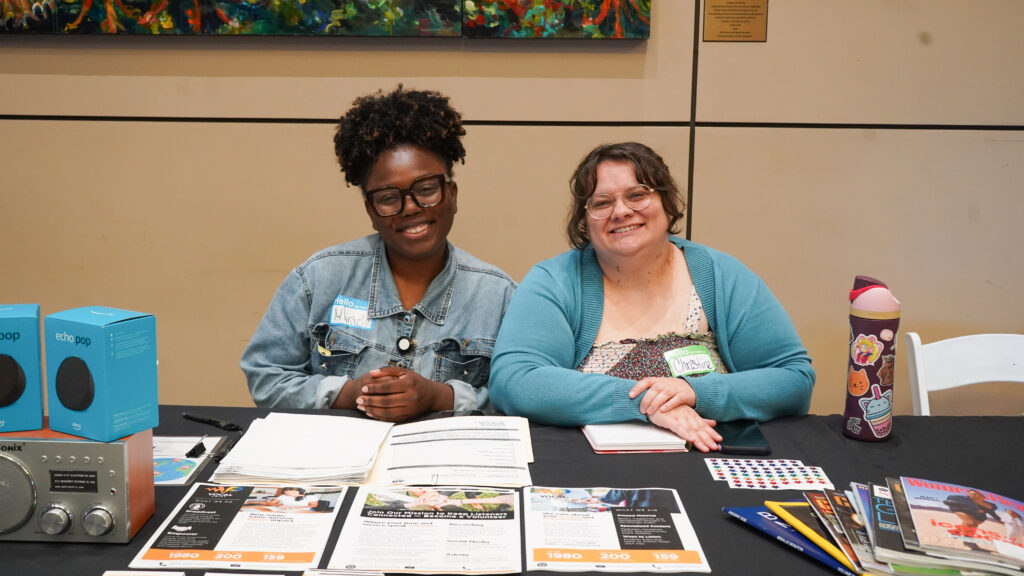 Two Women Sit Behind A Table At An Event, Smiling At The Camera. One Woman Has Curly Hair And Glasses, While The Other Has Shoulder-Length Hair And Is Wearing A Blue Cardigan. The Table Is Covered With Informational Materials, A Radio, And Two Blue Devices.