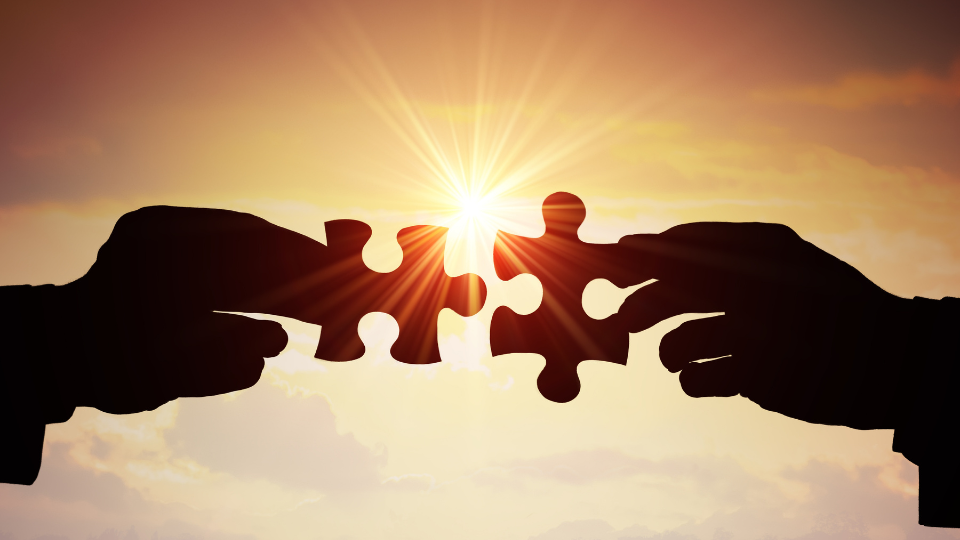 Two Hands Reaching Out To Connect Two Puzzle Pieces Against A Sunset Background, Symbolizing Collaboration And Unity.
